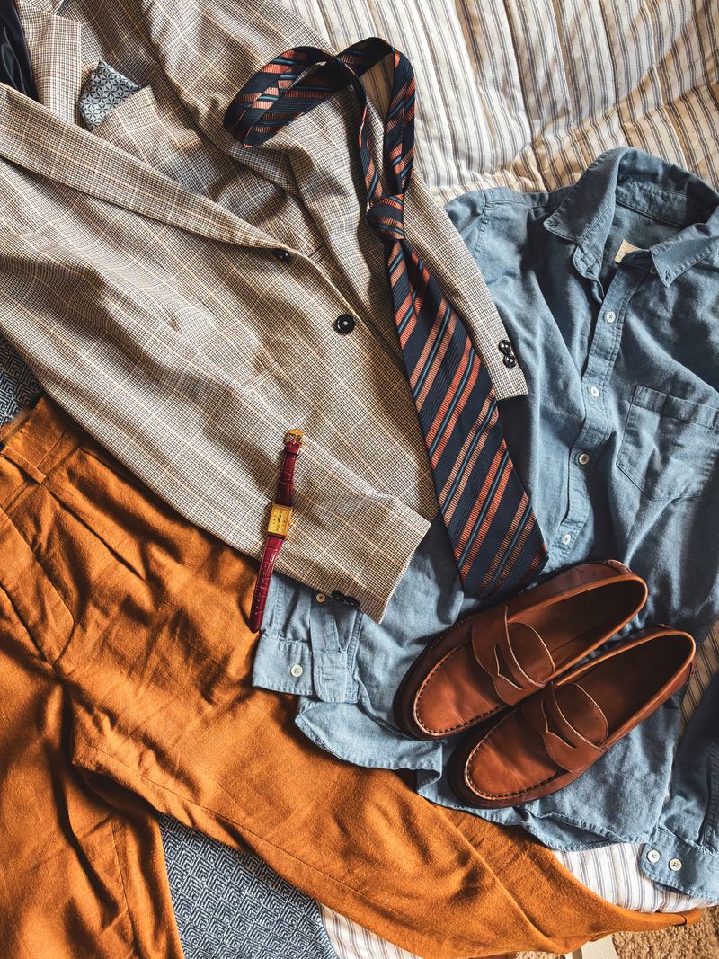 Flat lay of classic men's clothing including a suit jacket, tie, loafers, and watch.